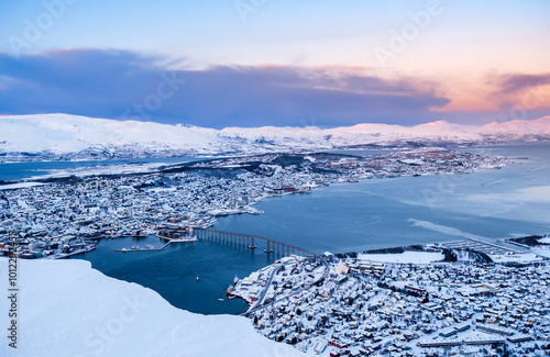 Aerial view of Tromso, Norway at sunset in winter from Storsteinen mountain. Tromsoya island with snowy roofs of Tromso downtown, harbor, Sandnessundet Bridge over Fjord and mountains in background