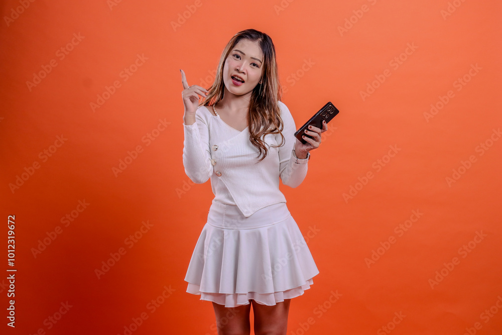 portrait of beautiful young asian female in white top standing shouting to camera holding mobile phone, cellphone and hand pointing on orange background isolation. for technology, promotion, lifestyle portrait of beautiful young asian female in white top standing shouting to camera holding mobile phone, cellphone and hand pointing on orange background isolation. for technology, promotion, lifestyle