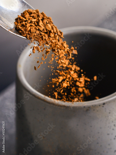 Grains of freeze dried instant coffee,falling from a silver tea spoon,into an empty cup,on a slate grey background..