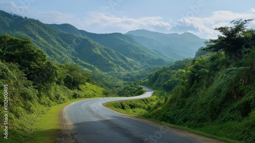 An asphalt road curving gently through lush green mountains, under a clear sky with light, wispy clouds on a sunny day.