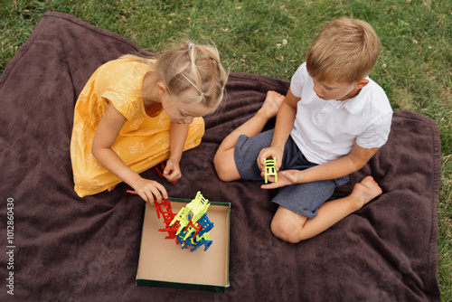 Two children seated on a blanket outdoors, engaging in play with colorful toys. The scene evokes a sense of fun and innocence.