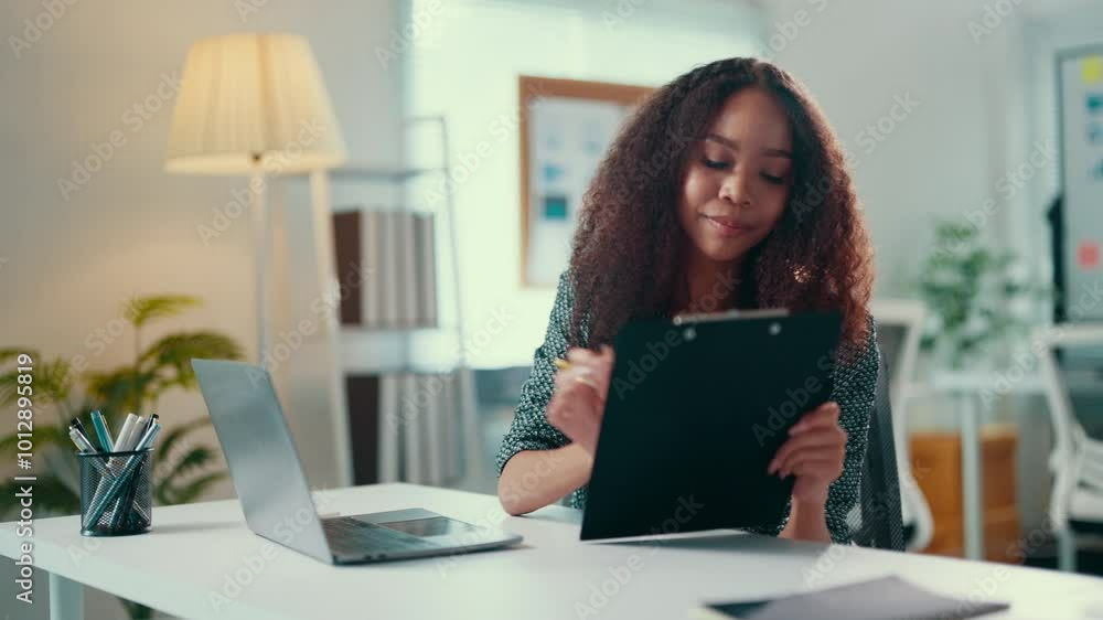 Young professional woman sits at her desk, taking notes on a clipboard while participating in a work video call, showcasing focus and engagement in a remote meeting setting Young professional woman sits at her desk, taking notes on a clipboard while participating in a work video call, showcasing focus and engagement in a remote meeting setting