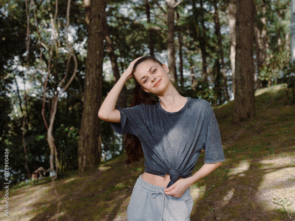 Cheerful young woman in casual clothing standing outdoors, touching her hair, surrounded by lush greenery Nature and carefree lifestyle concept