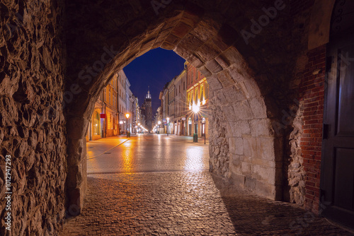 View through Florianska Gate in Krakow, Poland, showing the cobblestone street leading towards St. Mary's Basilica at night. The historic gate and street are illuminated with streetlights. photo