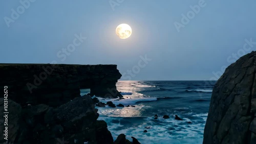 A glowing full moon rises over a rugged rocky coastline. Waves gently crash against the rocks reflecting the moon's silvery light in the calm twilight waters of the ocean.