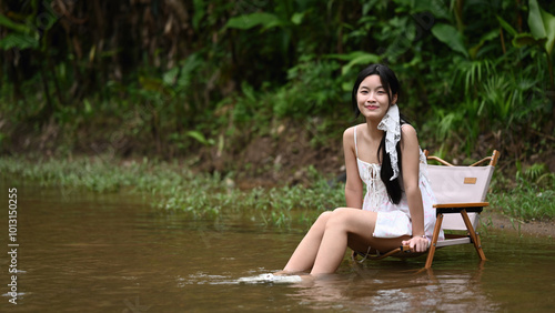 A teenage girl sits on a small chair, dipping her feet in a calm stream, smiling and enjoying a peaceful moment in nature