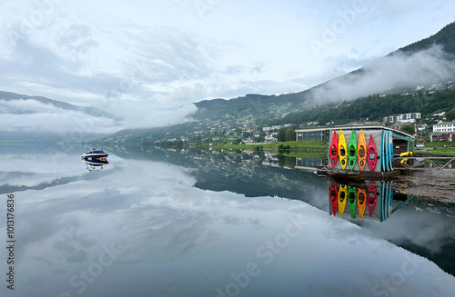 Colourful kayaks mirror reflection on peaceful mountain lake in misty morning, patches of snow.