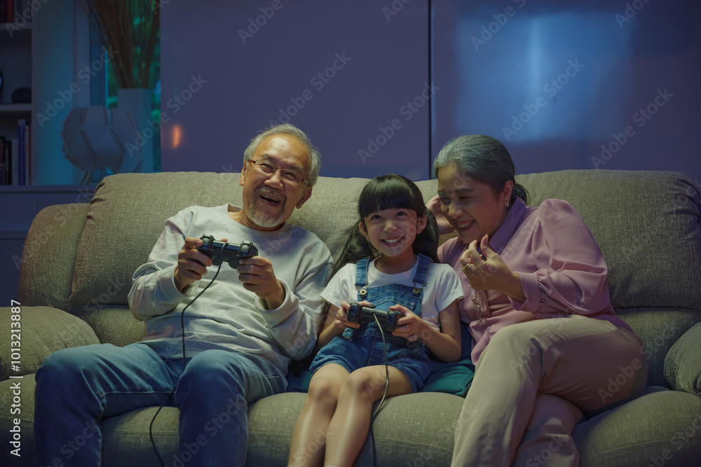 Happy Asian family bonding over a video game on a cozy couch, little girl playing video games console with senior grandparents together with controller joysticks while sitting on the sofa at night Happy Asian family bonding over a video game on a cozy couch, little girl playing video games console with senior grandparents together with controller joysticks while sitting on the sofa at night