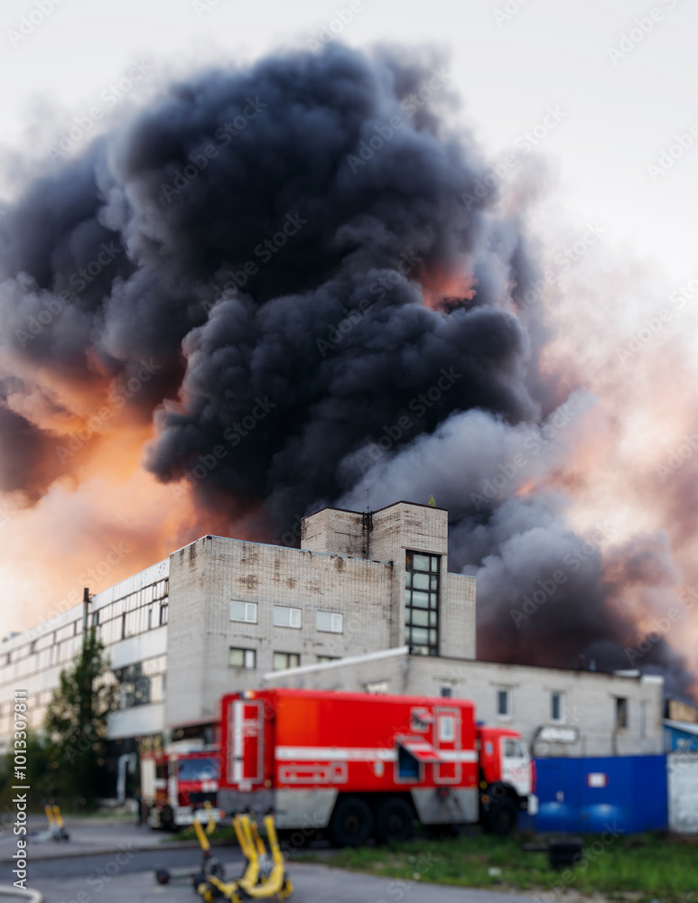 Fototapeta premium Massive large blaze fire in the city with large column plume of smoke, blazing warehouse factory, storage building is burning, putting out the fire, extinguish the fire, arson, burning house damage