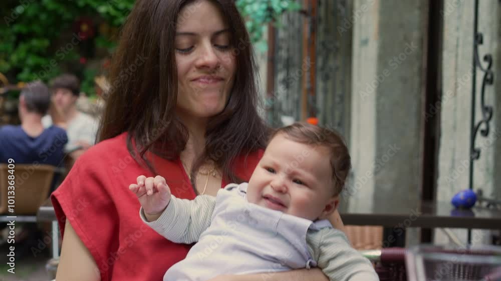 Mother holding baby, brushing her hair back while seated outdoors. The baby looks around curiously, showing interest in the surroundings as they share a calm and relaxed moment Mother holding baby, brushing her hair back while seated outdoors. The baby looks around curiously, showing interest in the surroundings as they share a calm and relaxed moment