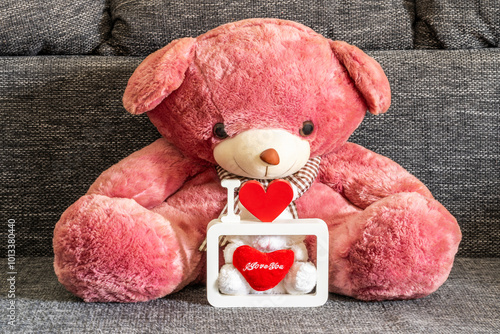 A large, plush teddy bear takes center stage, radiating a soft pink hue. In front of the bear, theres a small white box with a clear lid. Inside, a red heart shaped object bears the word I Love you. photo