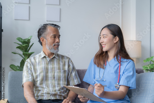 Asian female doctor wearing blue scrubs is sitting on a sofa, smiling and talking with a senior patient while holding a clipboard and pen