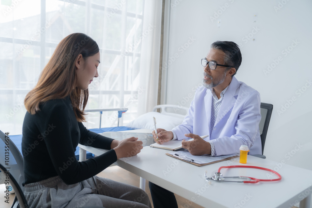 Fototapeta premium Senior male doctor is sitting at his desk and taking notes while discussing with a young female patient about her health in a hospital room