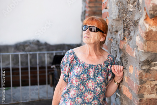 A beautiful older woman in sunglasses rests under an ancient stone wall, it's very hot outside, the woman has headache from stuffiness and heat. Health concept photo