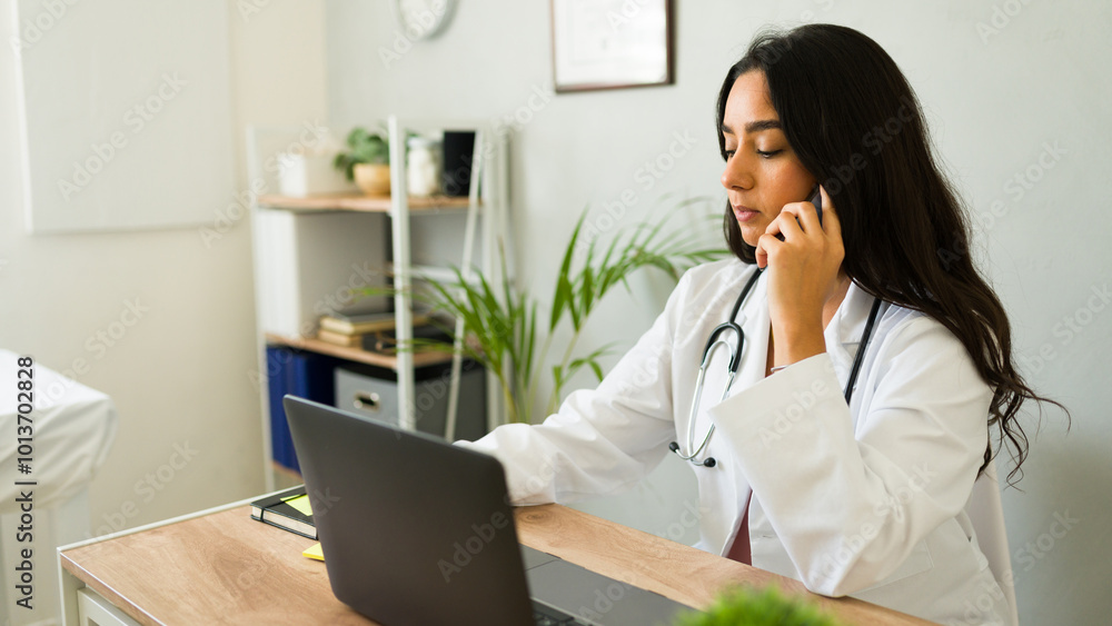 Woman physician is seated at her desk in her office, using her laptop and speaking on the phone Woman physician is seated at her desk in her office, using her laptop and speaking on the phone