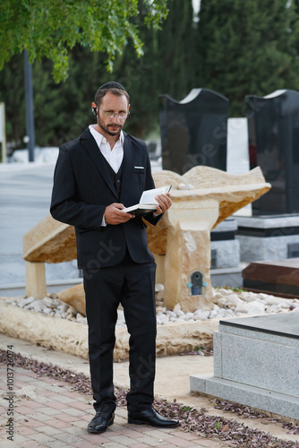 Jew male dressed in a formal black suit stands in a cemetery surrounded by gravestones. Jewish man with a beard, wearing a kippah, reading a prayer from the Torah. The prayer for the deceased, funeral