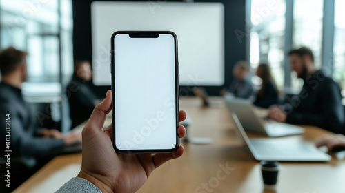 Obraz na płótnie A hand holding a mobile phone with a white screen in an office meeting room, the background showing a conference table, laptops, and colleagues in discussion, slightly blurred w salonie