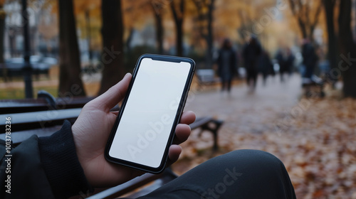 A close-up shot of a mobile phone with a white screen held by a person sitting on a park bench, with trees and people in the background, blurred for a sense of depth and motion