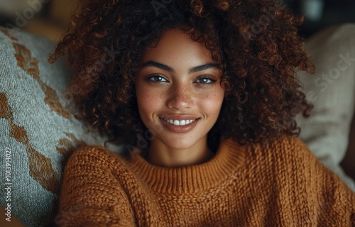 Smiling woman with curly hair wearing a cozy sweater, relaxing on a sofa in a warmly lit living room