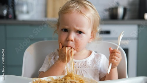 Joyful baby attempts to eat homemade pasta bolognese with a fork while nestled in a high chair in cozy kitchen setting
