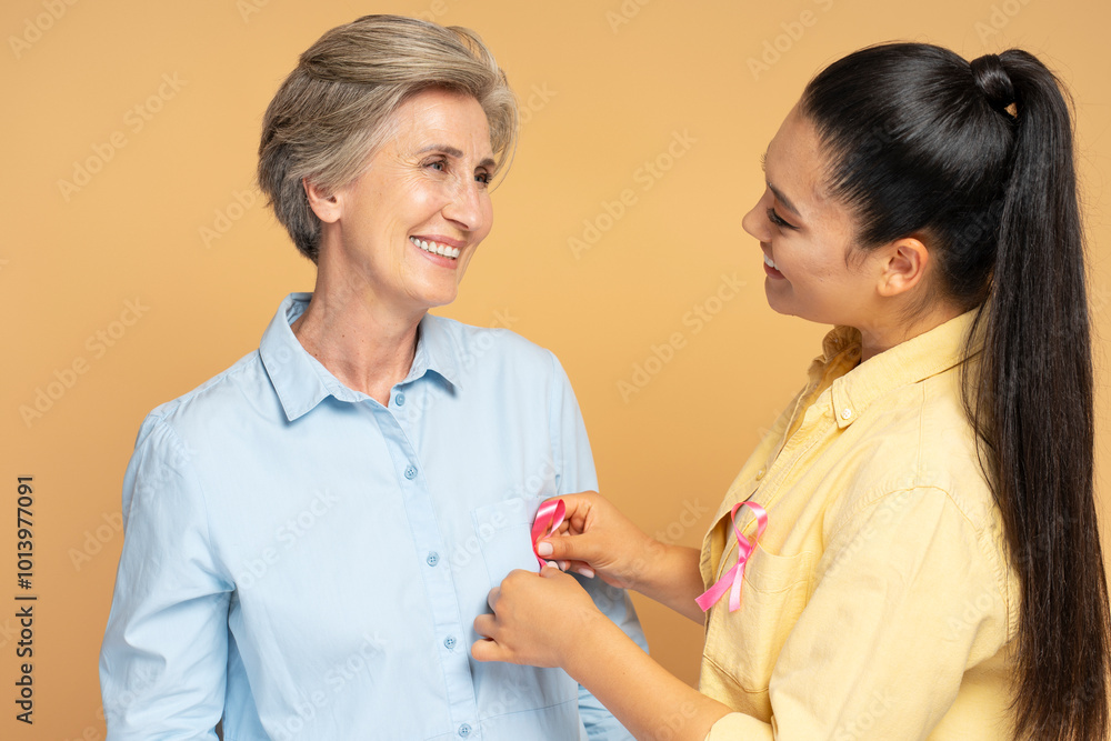 Young woman pinning pink ribbon on mature woman for breast cancer awareness Young woman pinning pink ribbon on mature woman for breast cancer awareness