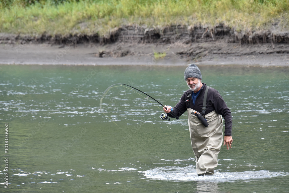 Older, attractive Caucasian man fishing, holding a fishing rod, wearing waders and standing in a river. Cool, autumn day in salmon season. Fishing rod is bent with a fish on the hook. Older, attractive Caucasian man fishing, holding a fishing rod, wearing waders and standing in a river. Cool, autumn day in salmon season. Fishing rod is bent with a fish on the hook.