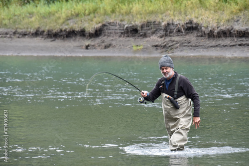 Older, attractive Caucasian man fishing, holding a fishing rod, wearing waders and standing in a river. Cool, autumn day in salmon season. Fishing rod is bent with a fish on the hook.