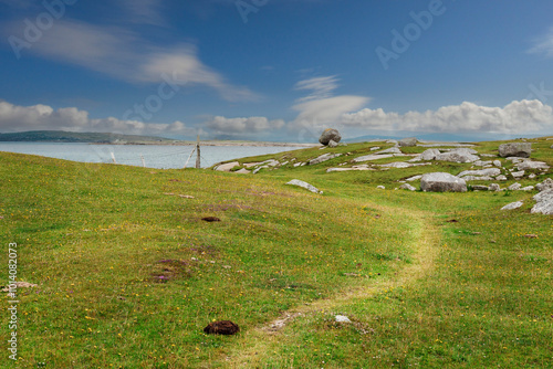 A grassy field with a path leading to a body of water. The sky is clear and blue, and there are some clouds in the background. Dog bay, county Galway, Ireland. Popular tourist area. photo