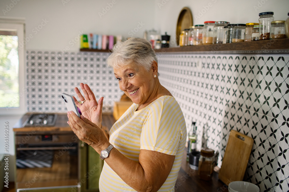 Happy senior woman waving at phone during video call in kitchen Happy senior woman waving at phone during video call in kitchen