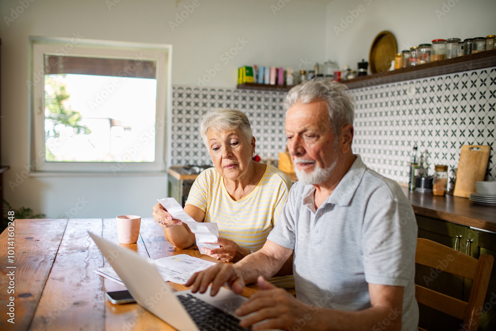 Elderly couple managing bills and finances on laptop in kitchen Elderly couple managing bills and finances on laptop in kitchen