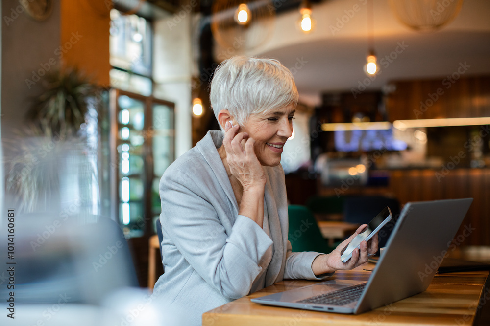 Senior businesswoman working on laptop in a cafe with wireless earbuds Senior businesswoman working on laptop in a cafe with wireless earbuds