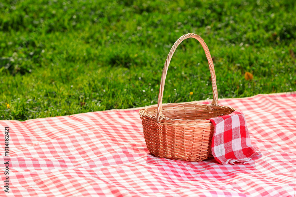 One picnic wicker basket with checkered napkin and blanket on green grass. Space for text One picnic wicker basket with checkered napkin and blanket on green grass. Space for text