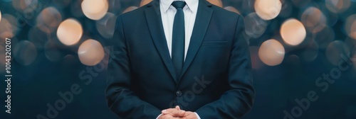 A businessman dressed in a formal black suit and tie stands with hands clasped in front, with a soft bokeh background of warm lights.