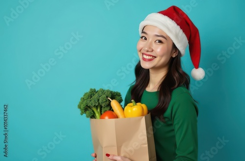 A cheerful young woman in a Santa hat carries a paper grocery bag filled with fresh vegetables, smiling against a solid blue background, conveying a festive and healthy holiday vibe