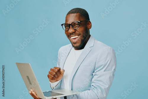 Smiling young man in stylish suit holding laptop, engaged in work with a joyful expression, set against a pastel blue background that conveys professionalism and approachability