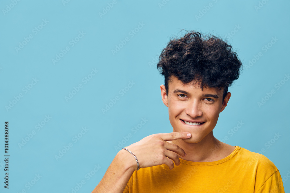 Young man with curly hair smiling playfully against a bright blue background He wears a yellow t shirt, exuding a cheerful and friendly vibe, inviting engagement and connection Young man with curly hair smiling playfully against a bright blue background He wears a yellow t shirt, exuding a cheerful and friendly vibe, inviting engagement and connection