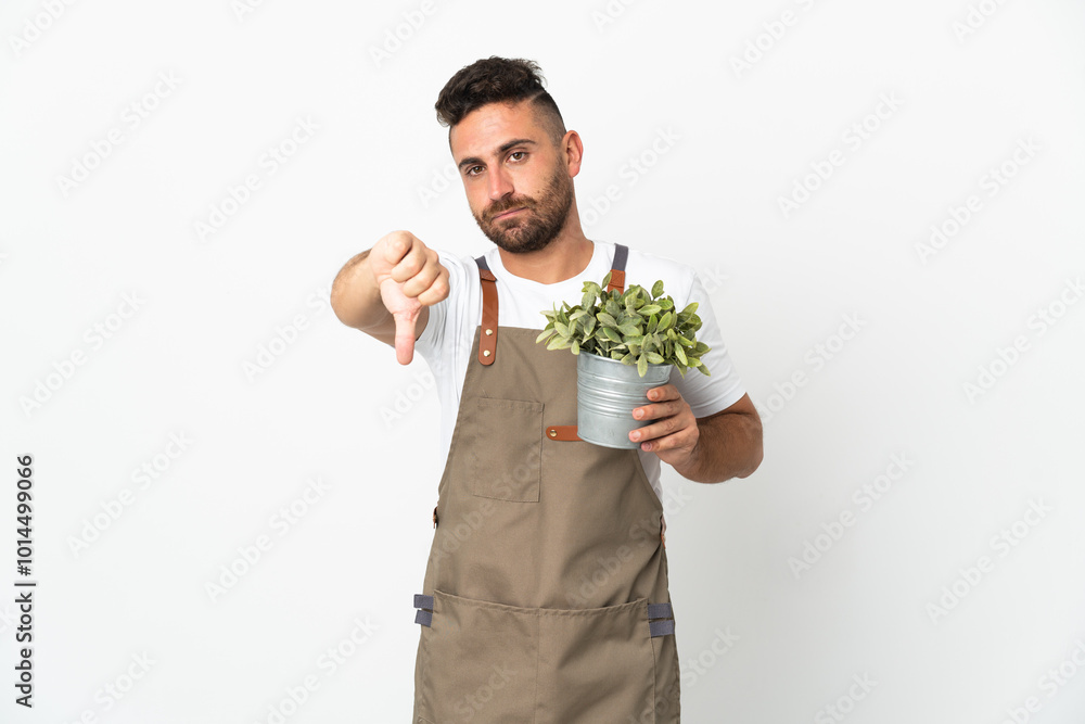 Gardener man holding a plant over isolated white background showing thumb down with negative expression Gardener man holding a plant over isolated white background showing thumb down with negative expression