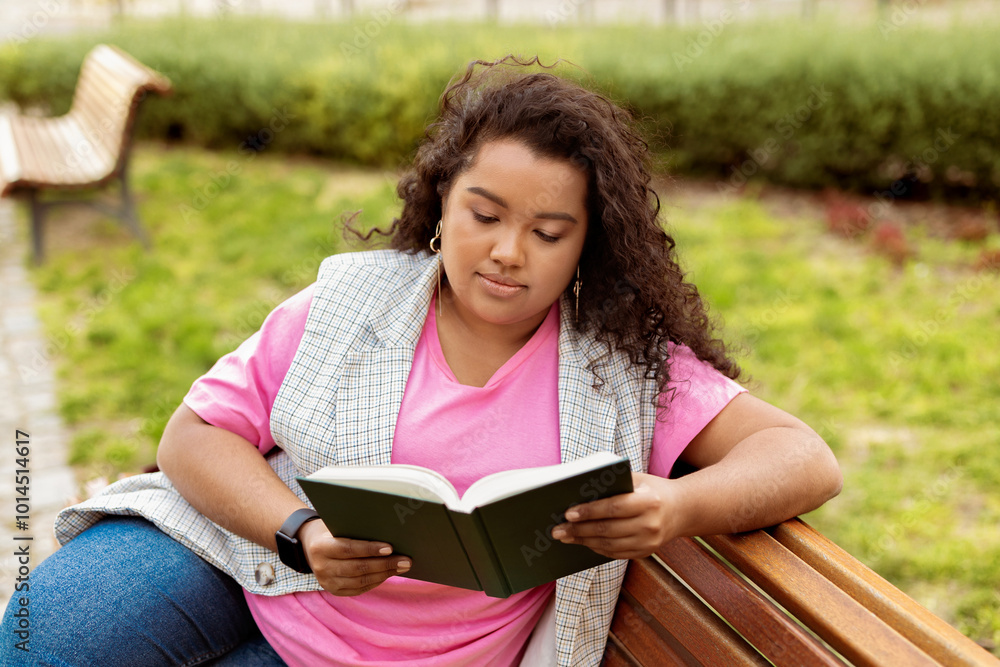Obraz premium A young woman sits on a wooden bench in a park, absorbed in reading a book. She enjoys the warmth of the sun while surrounded by green grass and flowering plants.