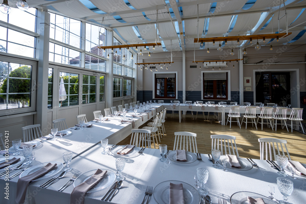 A bright, glass-walled banquet hall with tables covered with white tablecloths. Tableware ready to serve dishes. Photo taken during the day, with sunlight streaming in.