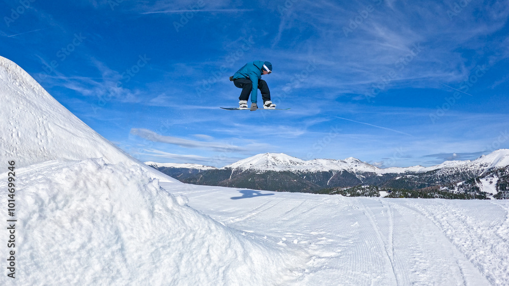Naklejka premium LOW ANGLE: Young male snowboarder rides off a kicker and does a cool grab trick. High-flying extreme snowboarder does a trick in the empty snow park. Active male tourist snowboarding in fun park.