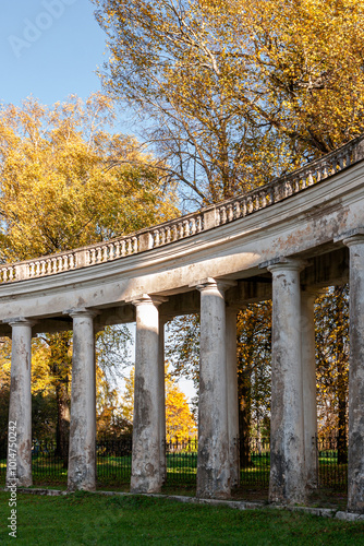 The image shows a neoclassical colonnade under restoration, with scaffolding surrounding the white columns. Trees with autumn leaves and a clear blue sky form the natural backdrop. photo