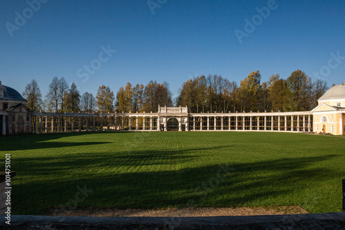 The image shows a neoclassical colonnade under restoration, with scaffolding surrounding the white columns. Trees with autumn leaves and a clear blue sky form the natural backdrop. photo