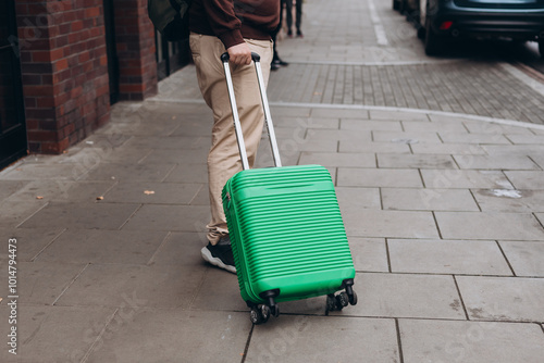 Unrecognizable man pulling green suitcase outdoors. Rear view. Copy space. Man walking on the street with travel bag. Male dragging baggage near hotel. Travel Fashion.
