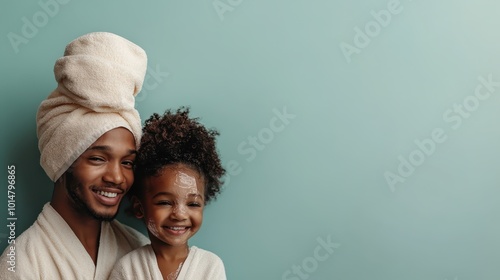 A father with a towel on his head smiles with his young daughter, both wearing robes, against a soft green background, suggesting a cozy spa moment together. photo