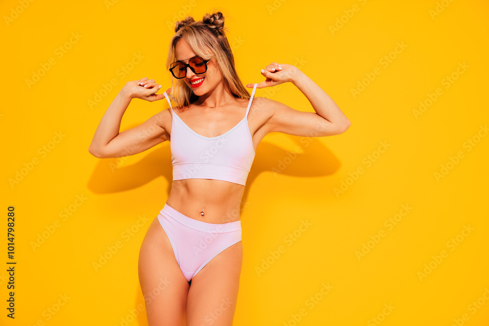 Young smiling blond model in summer swimwear colourful bathing suit. Sexy carefree woman having fun and going crazy. Female posing near yellow wall in studio. Cheerful and happy, sunglasses Young smiling blond model in summer swimwear colourful bathing suit. Sexy carefree woman having fun and going crazy. Female posing near yellow wall in studio. Cheerful and happy, sunglasses