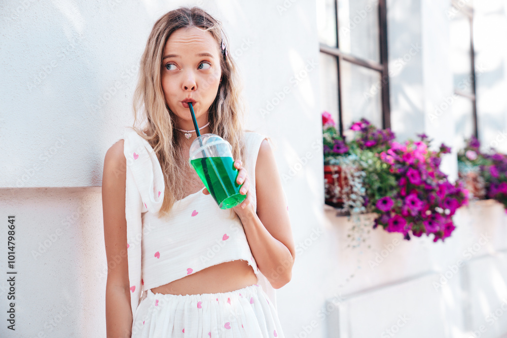 Young beautiful smiling hipster woman in trendy summer clothes. Sexy carefree model posing in the street. Holding and drinking fresh vegetable cocktail drink in plastic cup with straw. In sunny day Young beautiful smiling hipster woman in trendy summer clothes. Sexy carefree model posing in the street. Holding and drinking fresh vegetable cocktail drink in plastic cup with straw. In sunny day