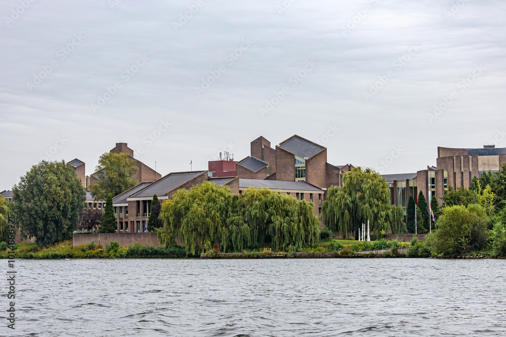 Obraz premium Gouvernement aan de Maas against grey sky, government building on river Meuse surrounded by leafy trees, seen from a boat, cloudy day in Maastricht, South Limburg, Netherlands