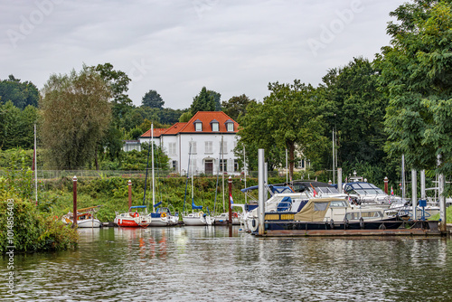 Small harbour with sailing boats and yachts anchored at quayside of on bank of Meuse river, white house surrounded leafy trees against grey sky, cloudy day in Maastricht, South Limburg, Netherlands photo