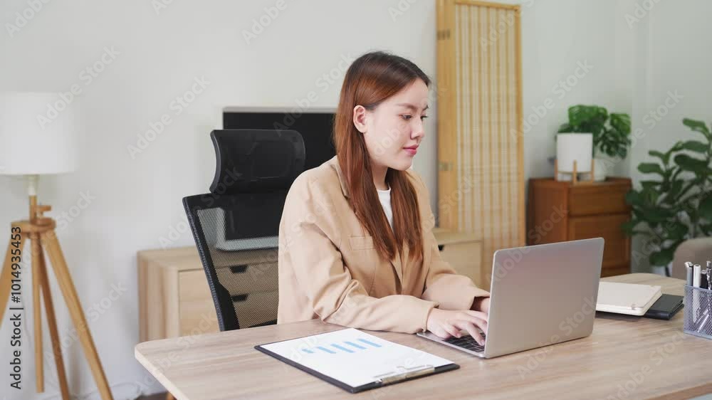 A young woman is sitting at her desk in a modern office, focused on her laptop. She is working in a clean and organized environment, with notebooks and office supplies around her A young woman is sitting at her desk in a modern office, focused on her laptop. She is working in a clean and organized environment, with notebooks and office supplies around her