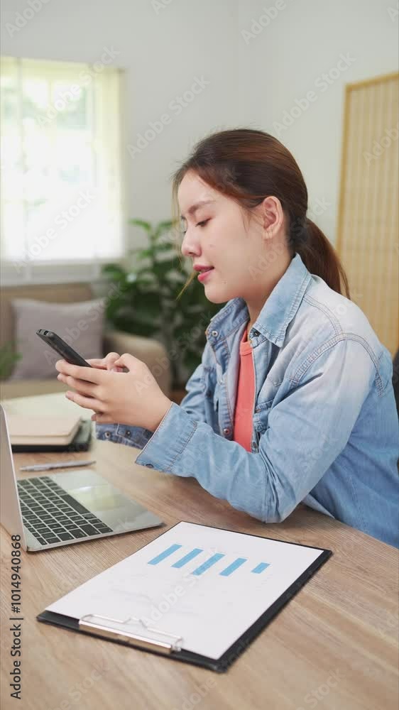 A young woman is smiling while looking at her phone in a modern office setting. She appears relaxed and cheerful, sitting at her desk with a laptop nearby in a bright, comfortable workspac